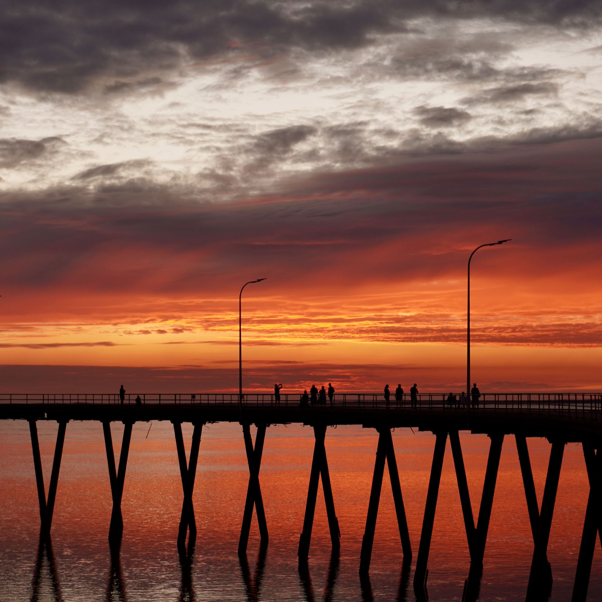 A jetty stretches out over the Indian Ocean at sunset with the silhouettes of pedestrians against the orange sky.