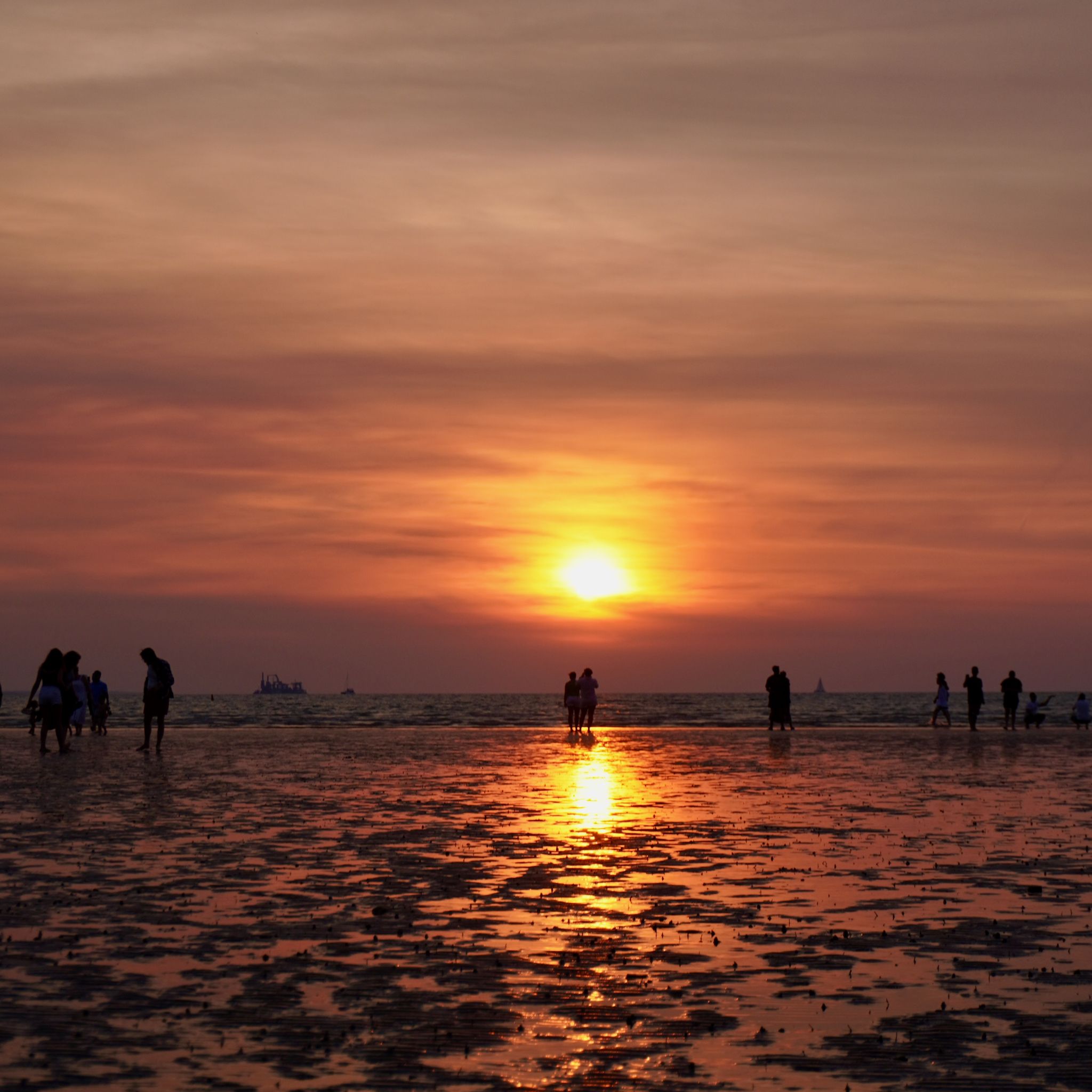 The sun sets over the water at Mindil Beach, creating a reflection in the low tide while tourists take pictures in the background.