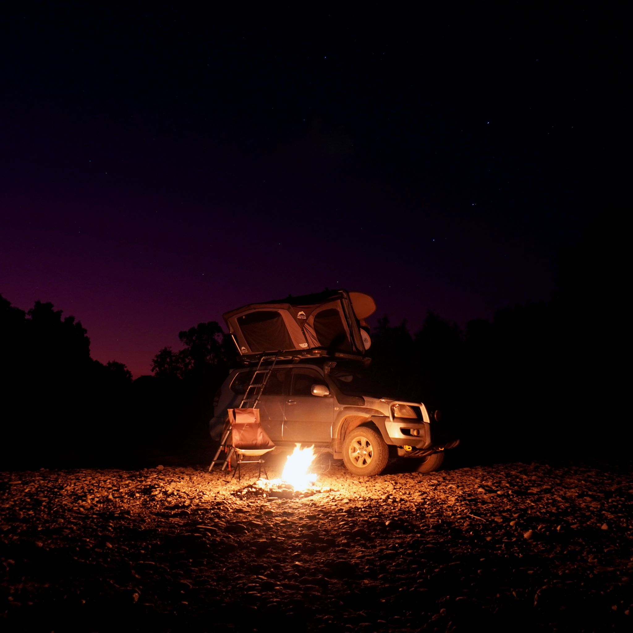 A campfire burning in front of Toyota Land Cruiser Prado with a rooftop tent opened and the starry sky above.