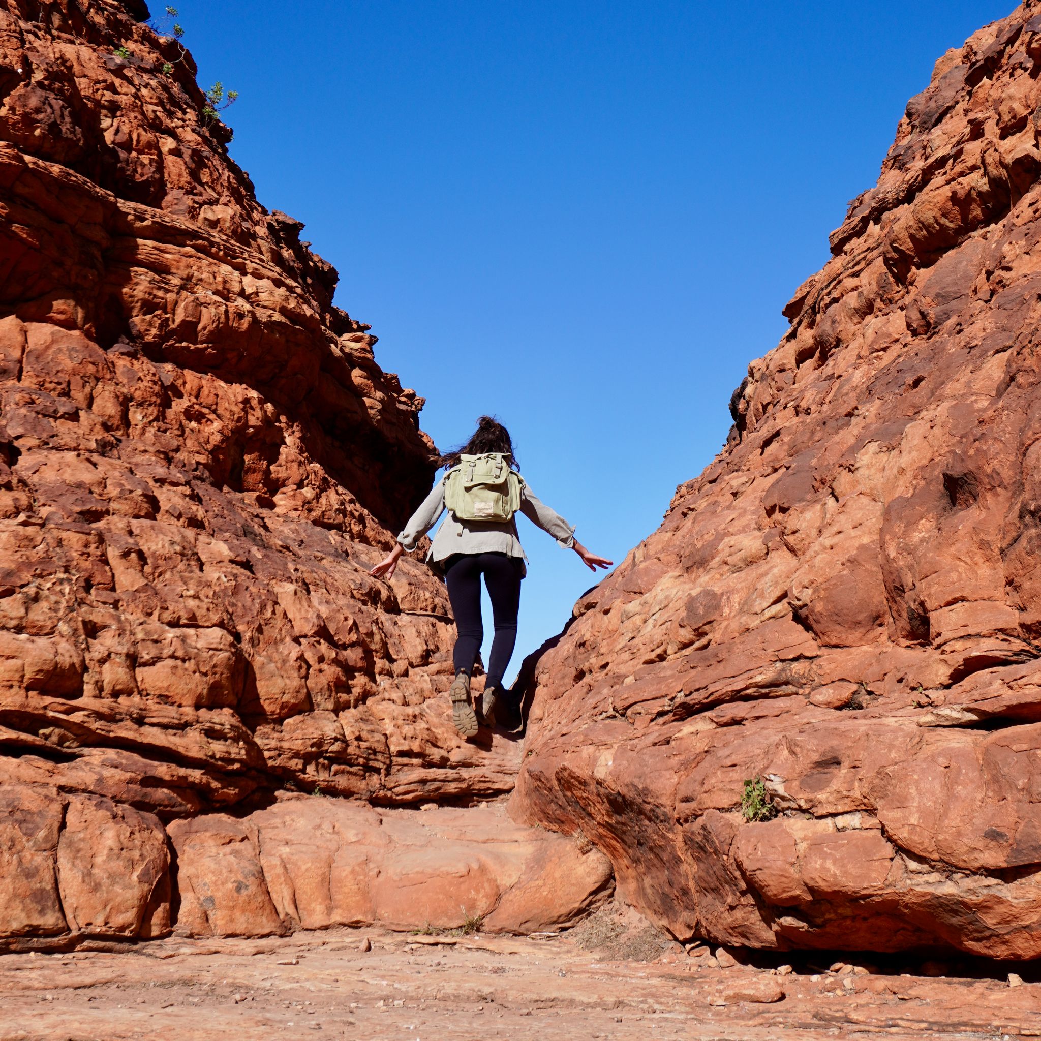 A person with a backpack jumps between red rock formations under a clear blue sky, typical of Australia's stunning landscapes.