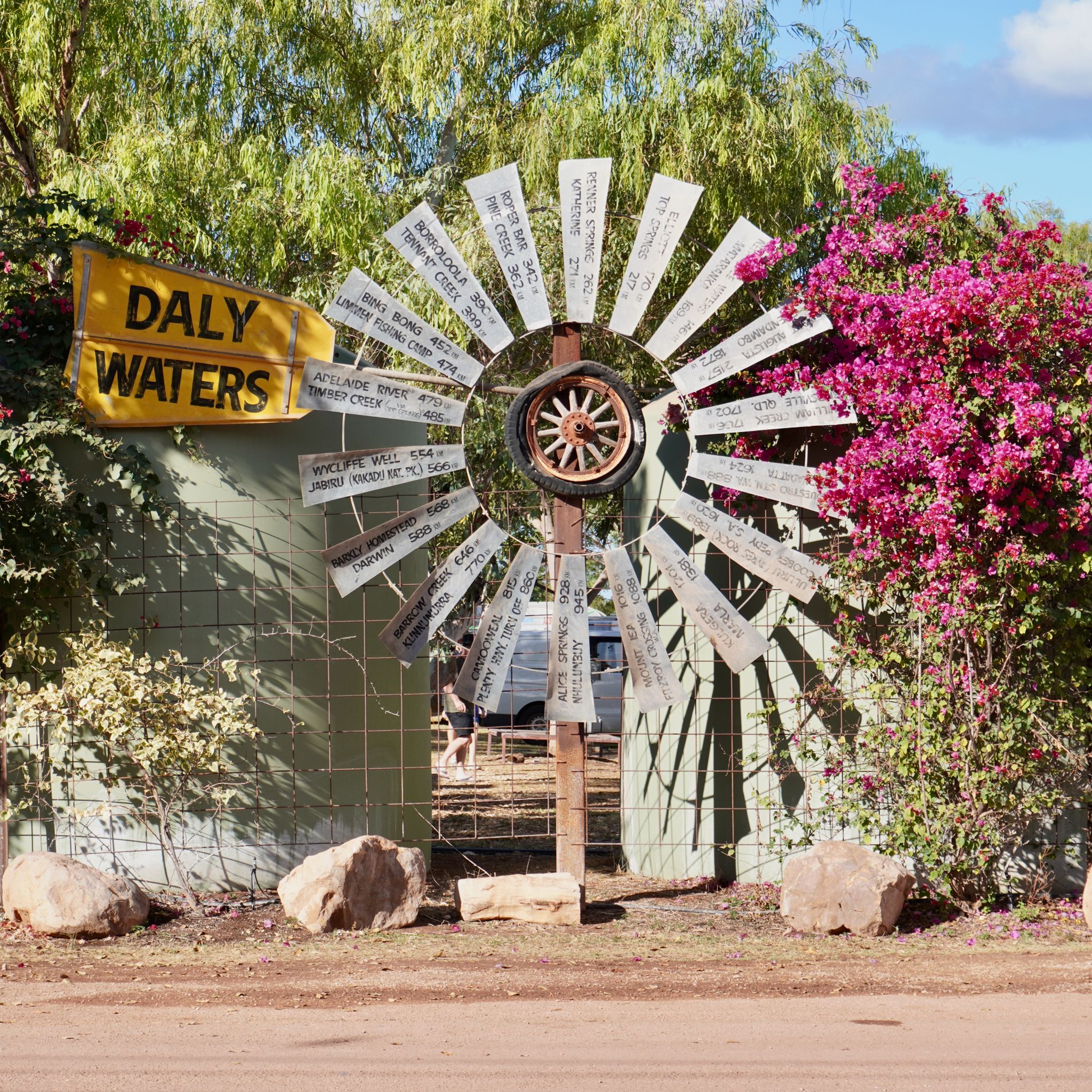 A metal windmill, with the distance to other 'nearby' towns painted on the blades. The windmill stands amidst pink flowers and a bright yellow 'Daly Waters' sign hangs beside it.